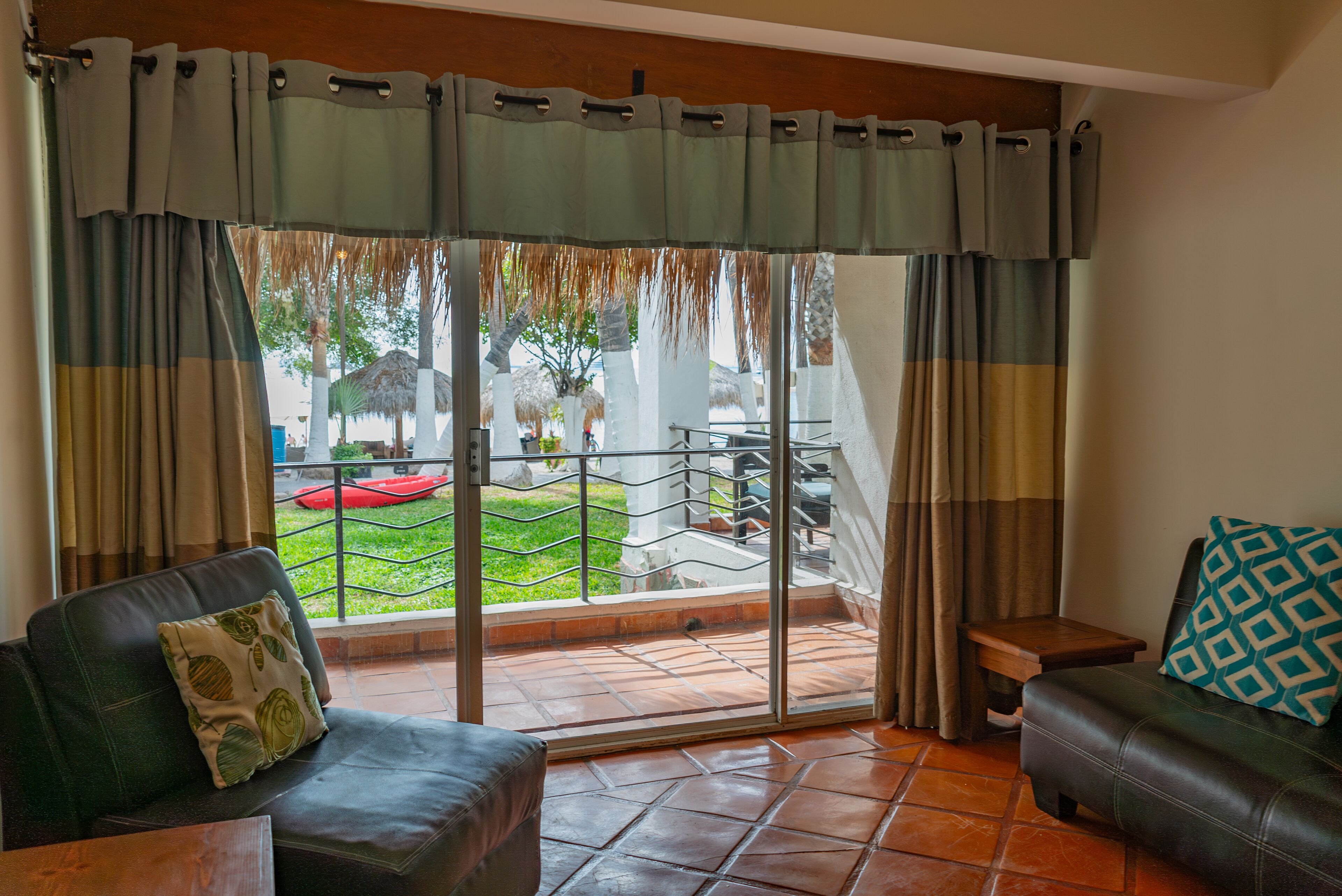 A room with a sliding glass door leading to a patio with a red chair and a thatched umbrella. There are two sofas with cushions, one with a patterned pillow and another with a geometric pillow.