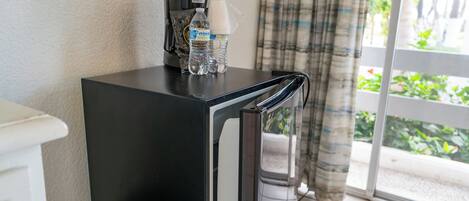 Un refrigerador negro compacto con una cafetera y una botella de agua encima, ubicado cerca de una puerta corrediza de vidrio que ofrece vistas al exterior.