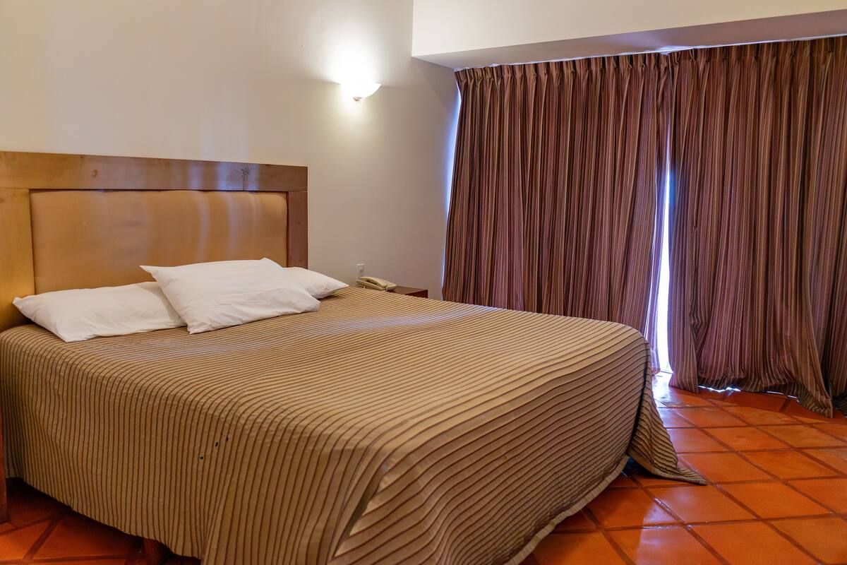 A hotel room with a large bed, a wooden headboard, a bedside table, a telephone, and a window with curtains.