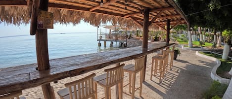 A beachside bar area with wooden chairs and a thatched roof.