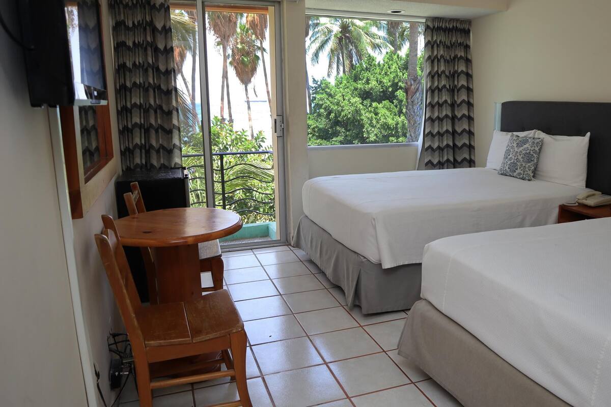 A hotel room with two beds, a dining table, chairs, a TV, and a view of palm trees.