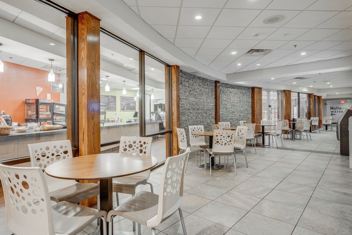 Un área de comedor interior moderna con mesas redondas y sillas perforadas, ventanas amplias y una pared de acento de piedra.