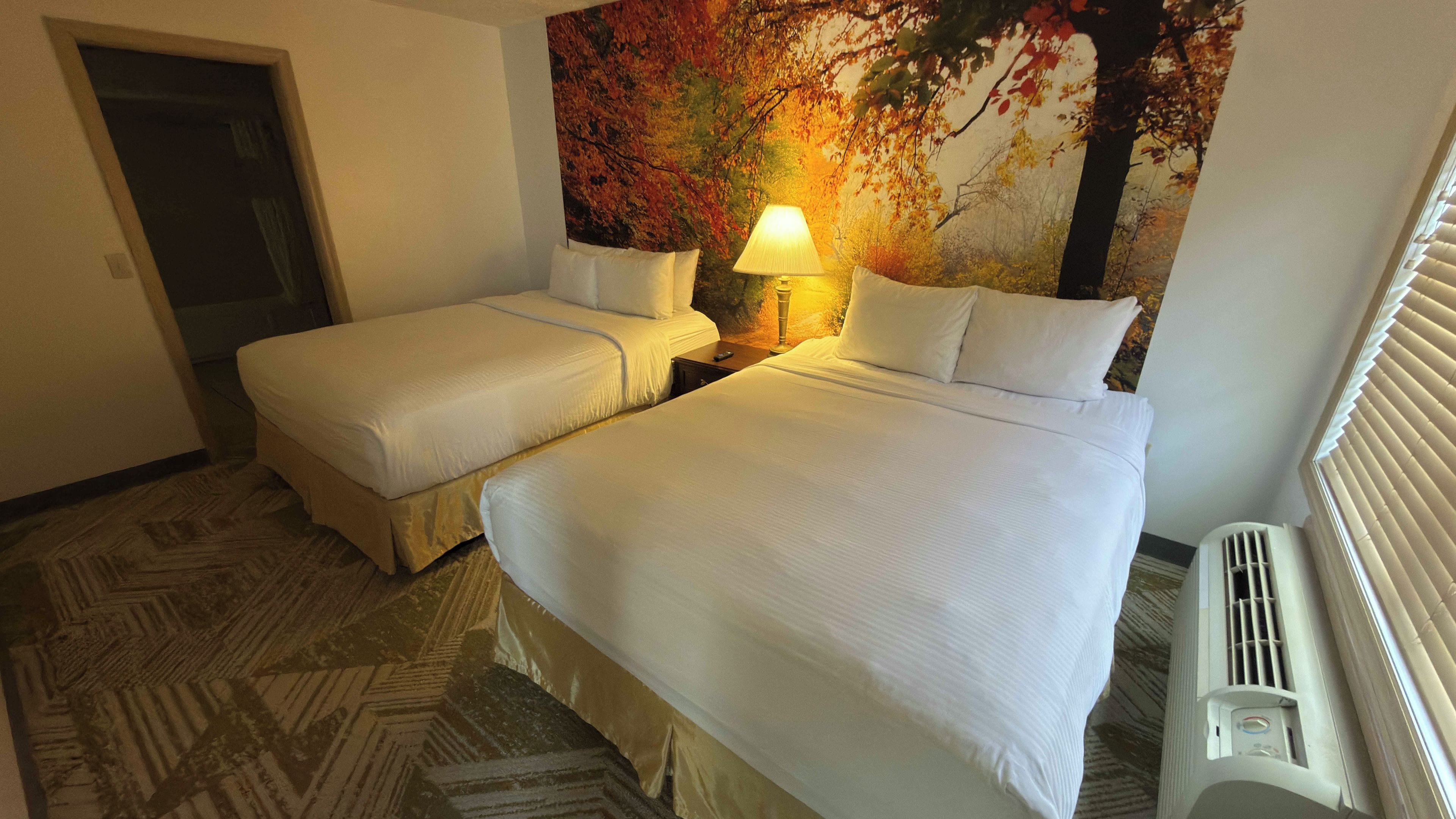Habitación de hotel con dos camas, una mesita de noche con lámpara, un mural en la pared y un radiador.