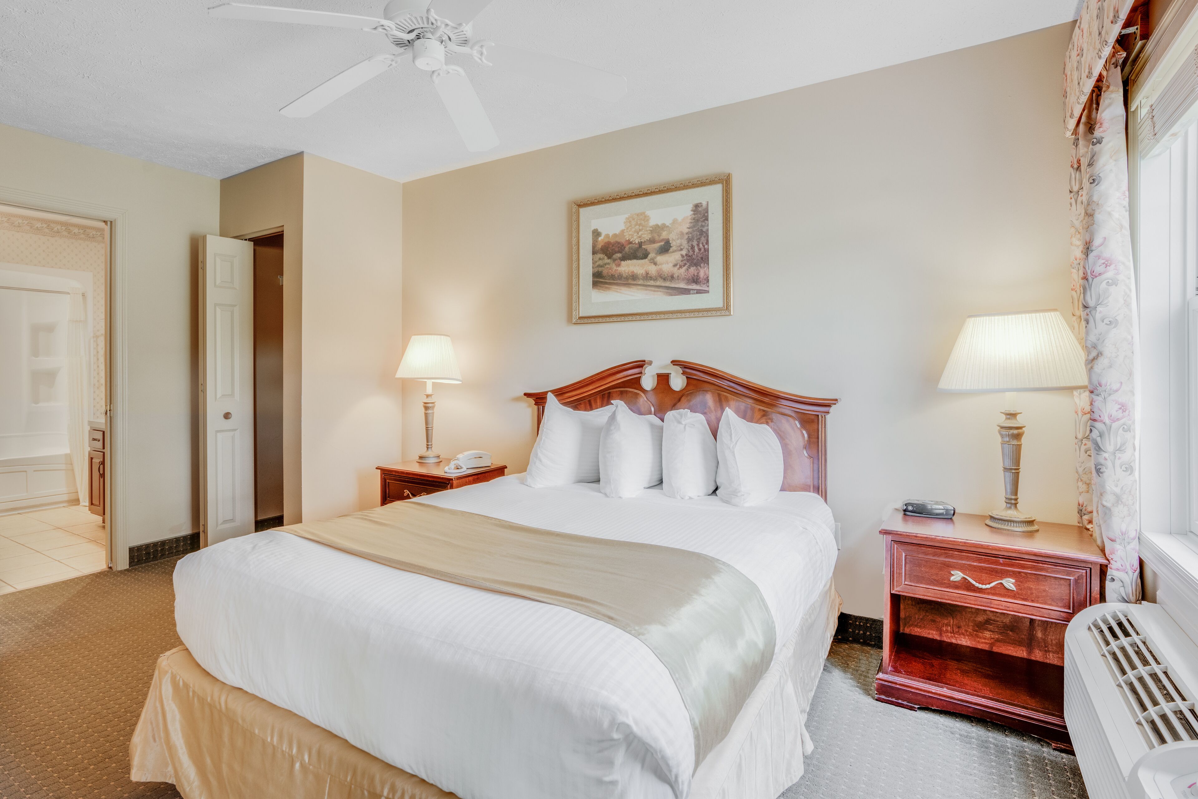 A hotel room with a large bed, a nightstand with a lamp, a painting on the wall, and a window with curtains.