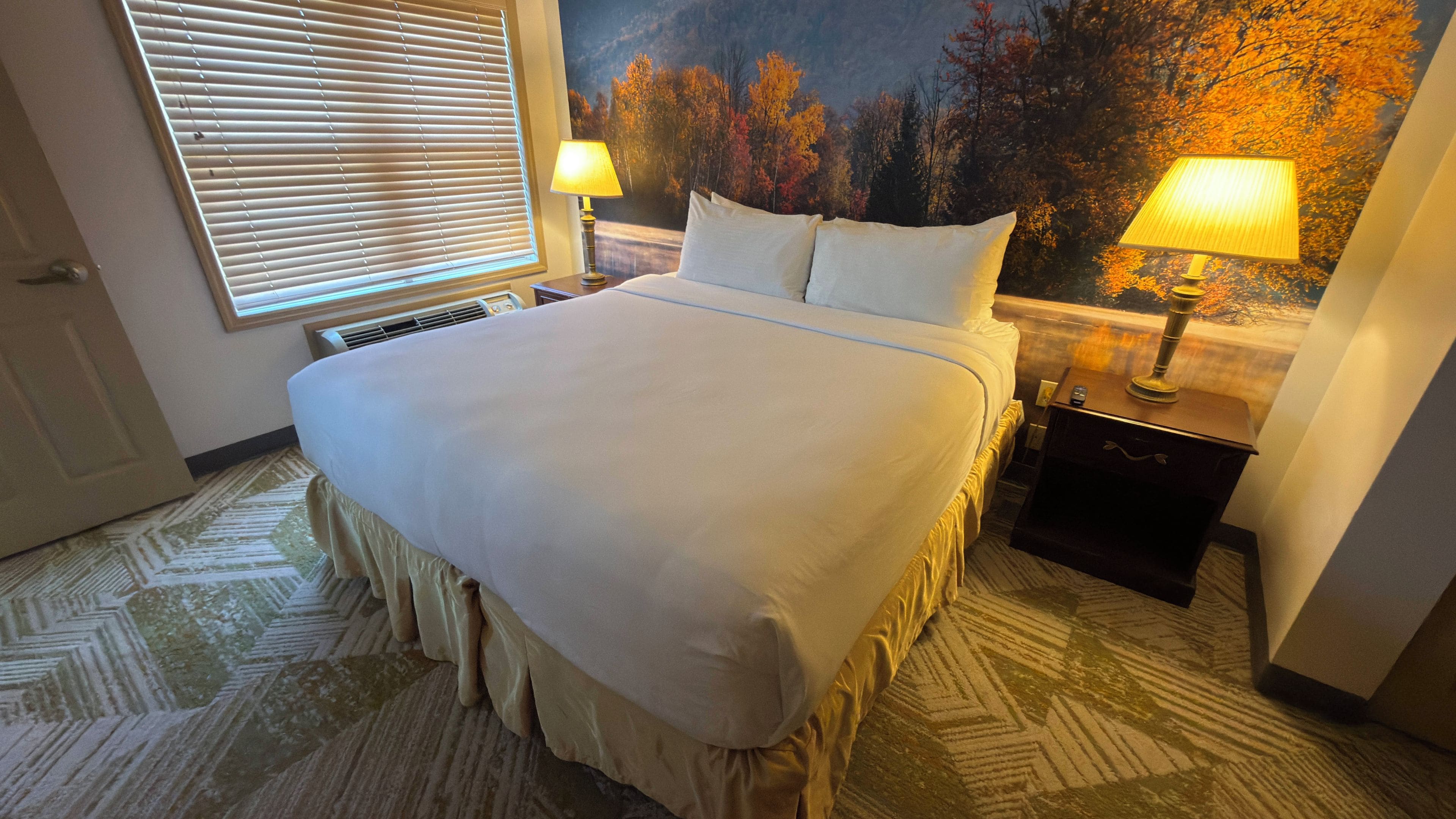 A hotel room with a large bed, two bedside lamps, a painting on the wall, and a patterned carpet.