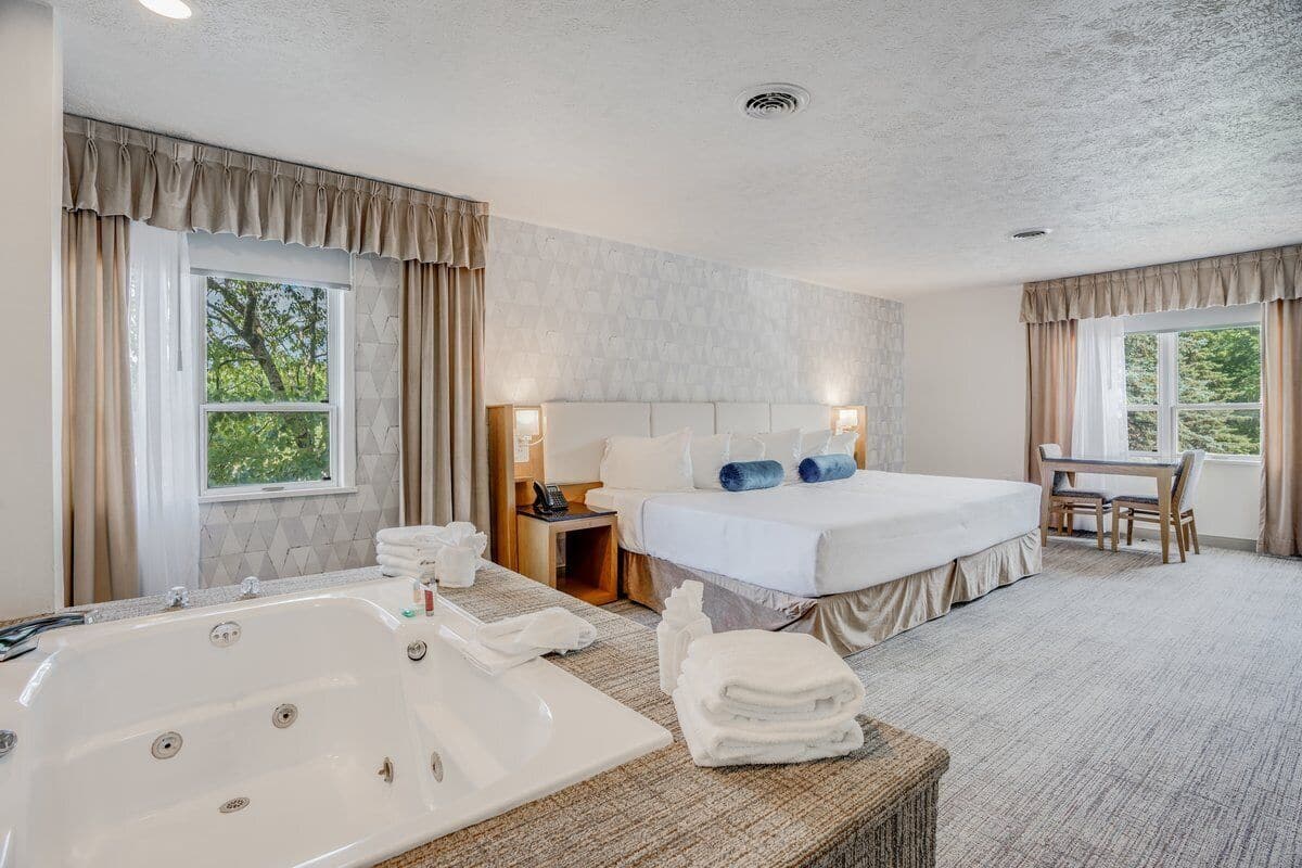 A hotel room with a large bed, a sitting area, a bathtub, and two windows with curtains.