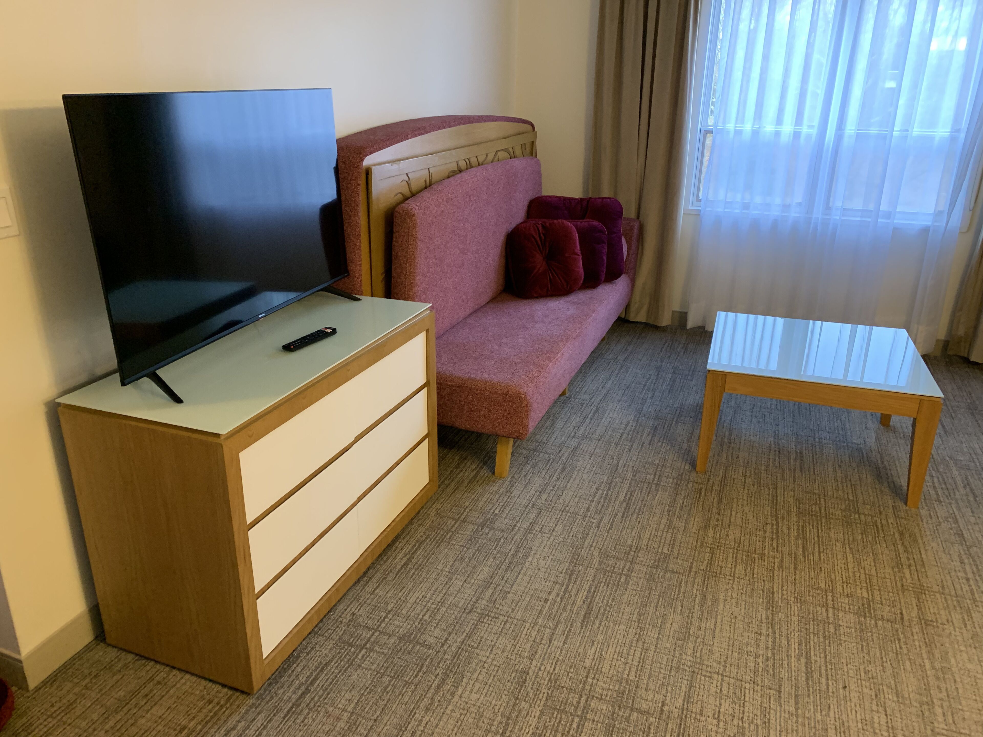A hotel room with a flat-screen TV on a wooden dresser, a pink sofa with red cushions, and a small wooden table.