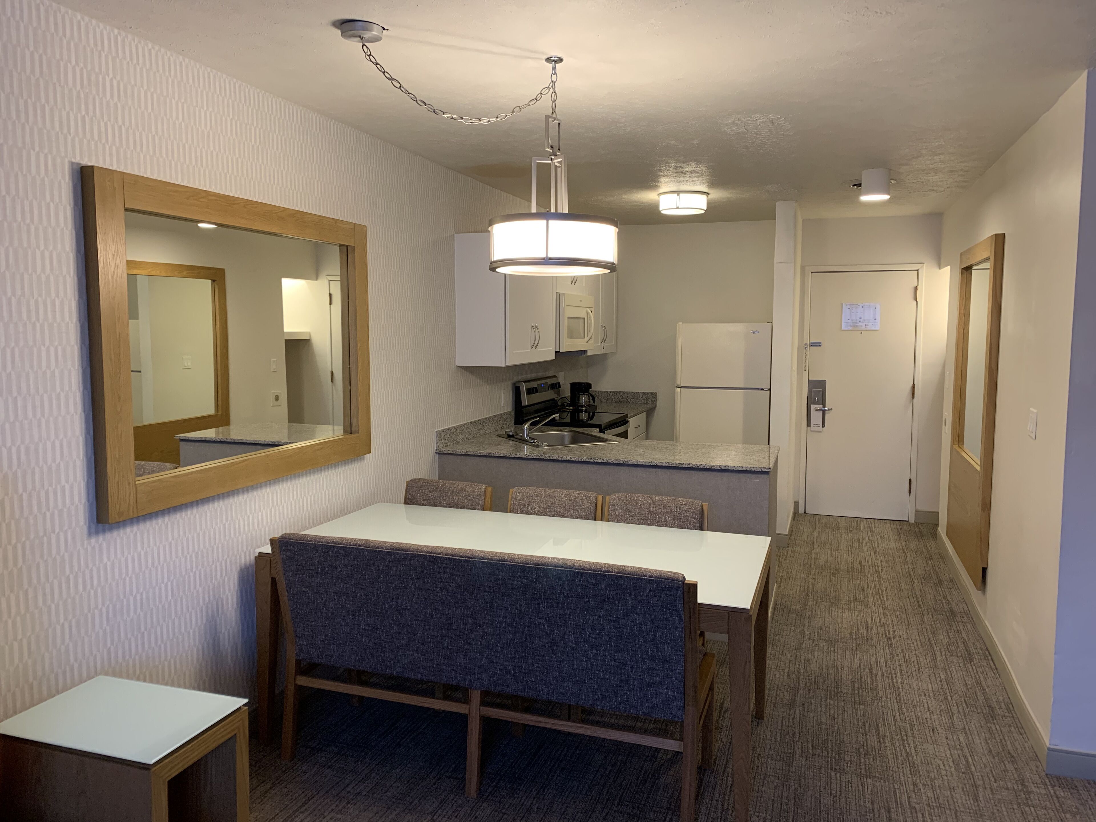 Ein kompaktes Hotelzimmer mit Küchenzeile, einer Essecke mit Tisch und Stühlen, einem großen Spiegel und einem Flur, der zu einer Tür führt.