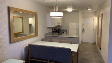 Habitación de hotel compacta con kitchenette, comedor con mesa y sillas, un espejo grande y un pasillo que conduce a una puerta.