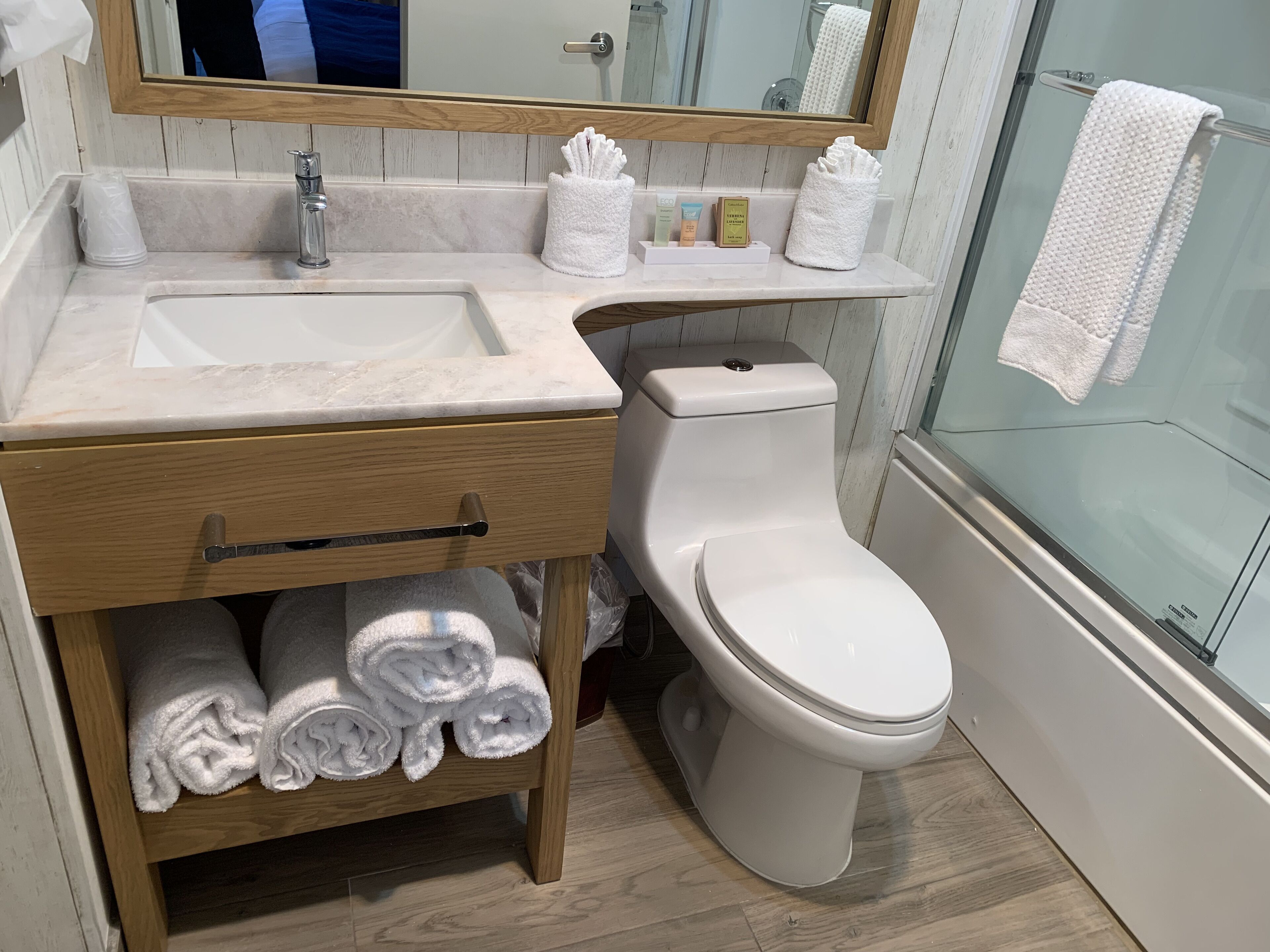 Ein Badezimmer mit einem weißen Waschbecken, einer Toilette und einem Spiegel. Auf dem Waschbecken liegen Handtücher und Toilettenartikel.