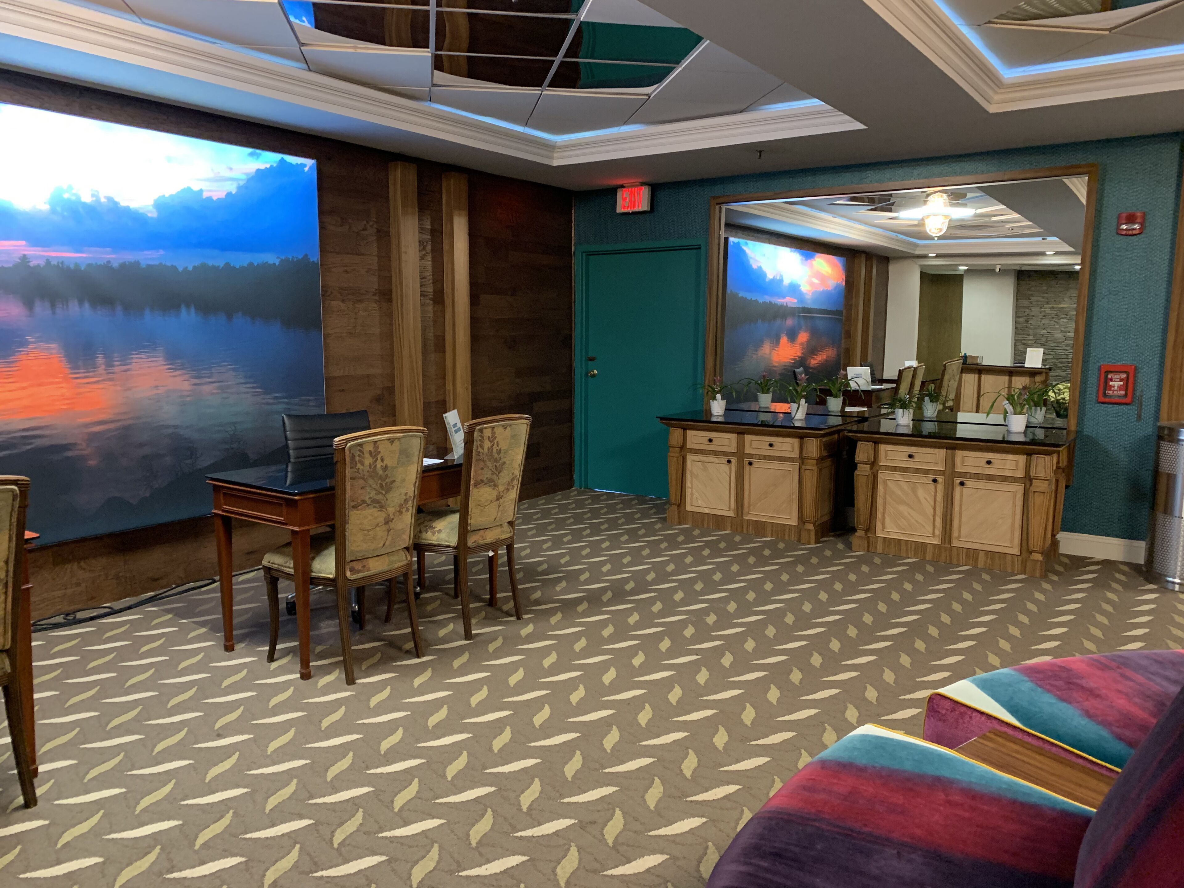 Une chambre d’hôtel comprenant un coin repas, une grande peinture représentant un coucher de soleil sur un lac, un coin cuisine et un canapé coloré.