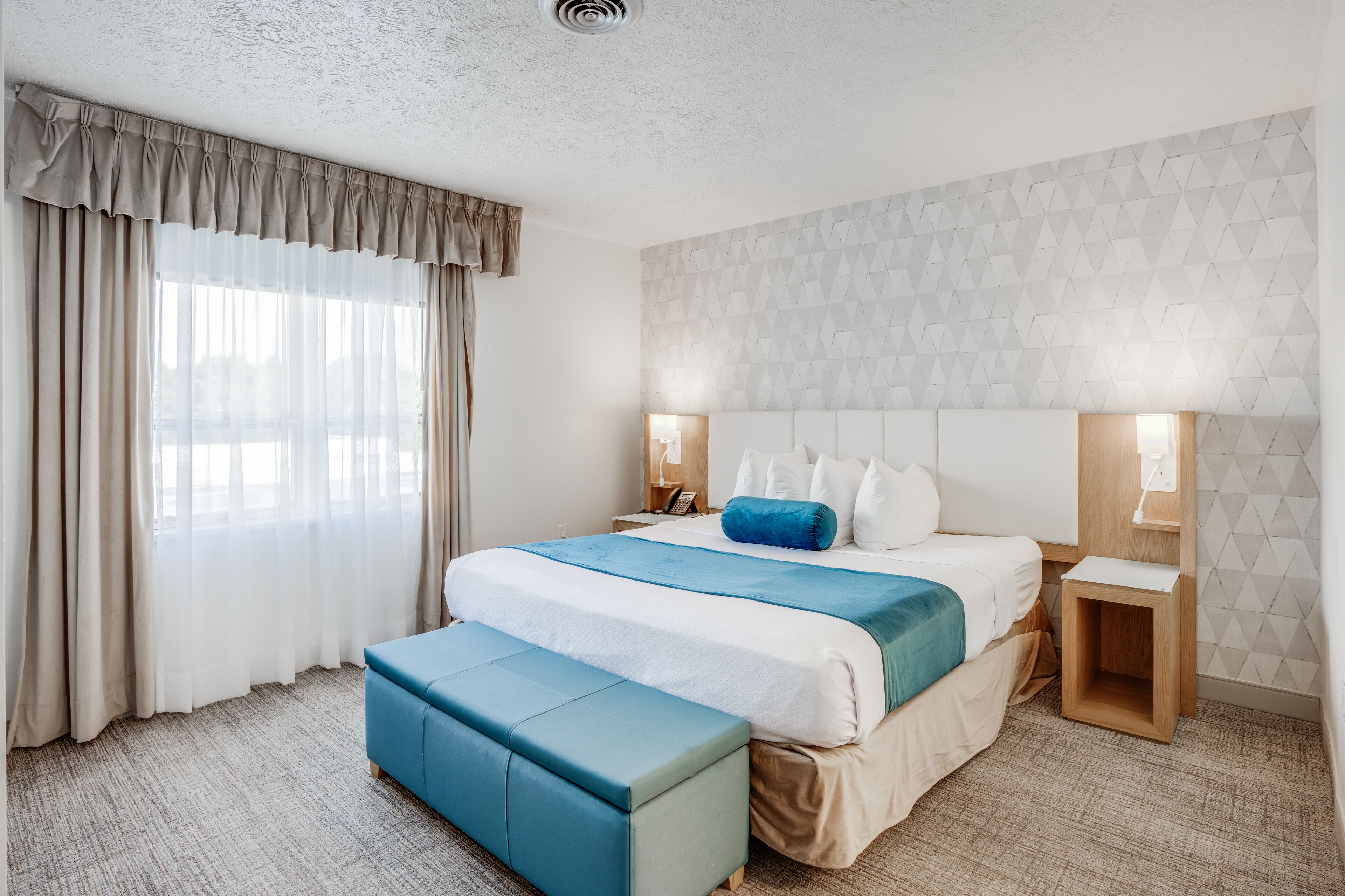 A hotel room with a large bed, a bedside table, a nightstand, a bench, and a geometric-patterned wall.