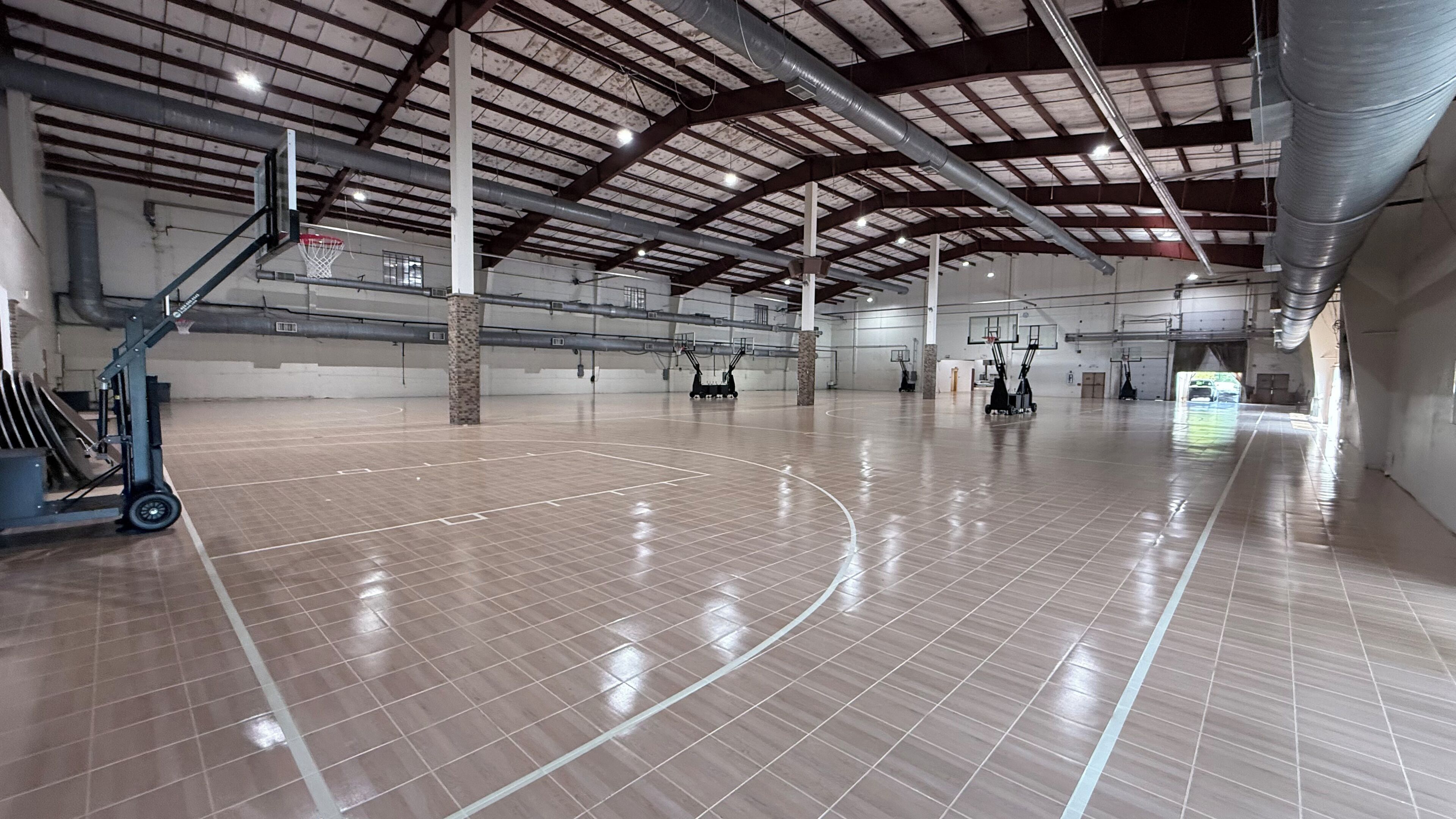 An indoor basketball court with a polished wooden floor, metal beams, and a high ceiling.