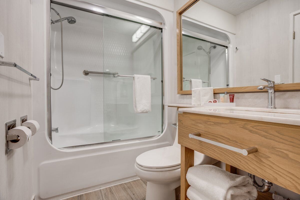 Un baño moderno con ducha de vidrio, inodoro blanco, tocador de madera con espejo y un perchero para toallas.