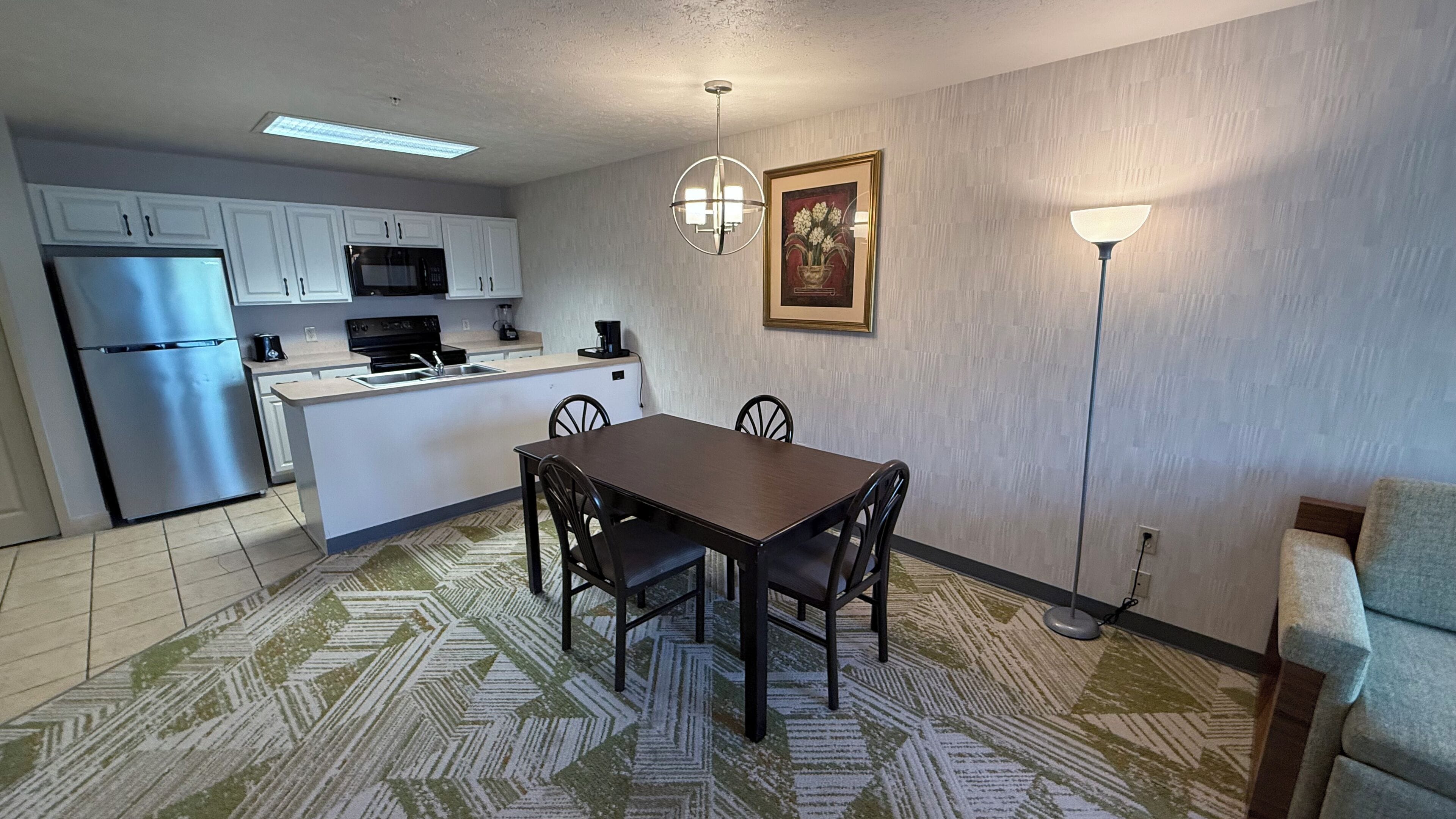 A compact kitchen with white cabinets, a stainless steel refrigerator, a microwave, and a coffee maker. There is a dining table with four chairs and a patterned rug. A floor lamp and a framed picture are on the wall.