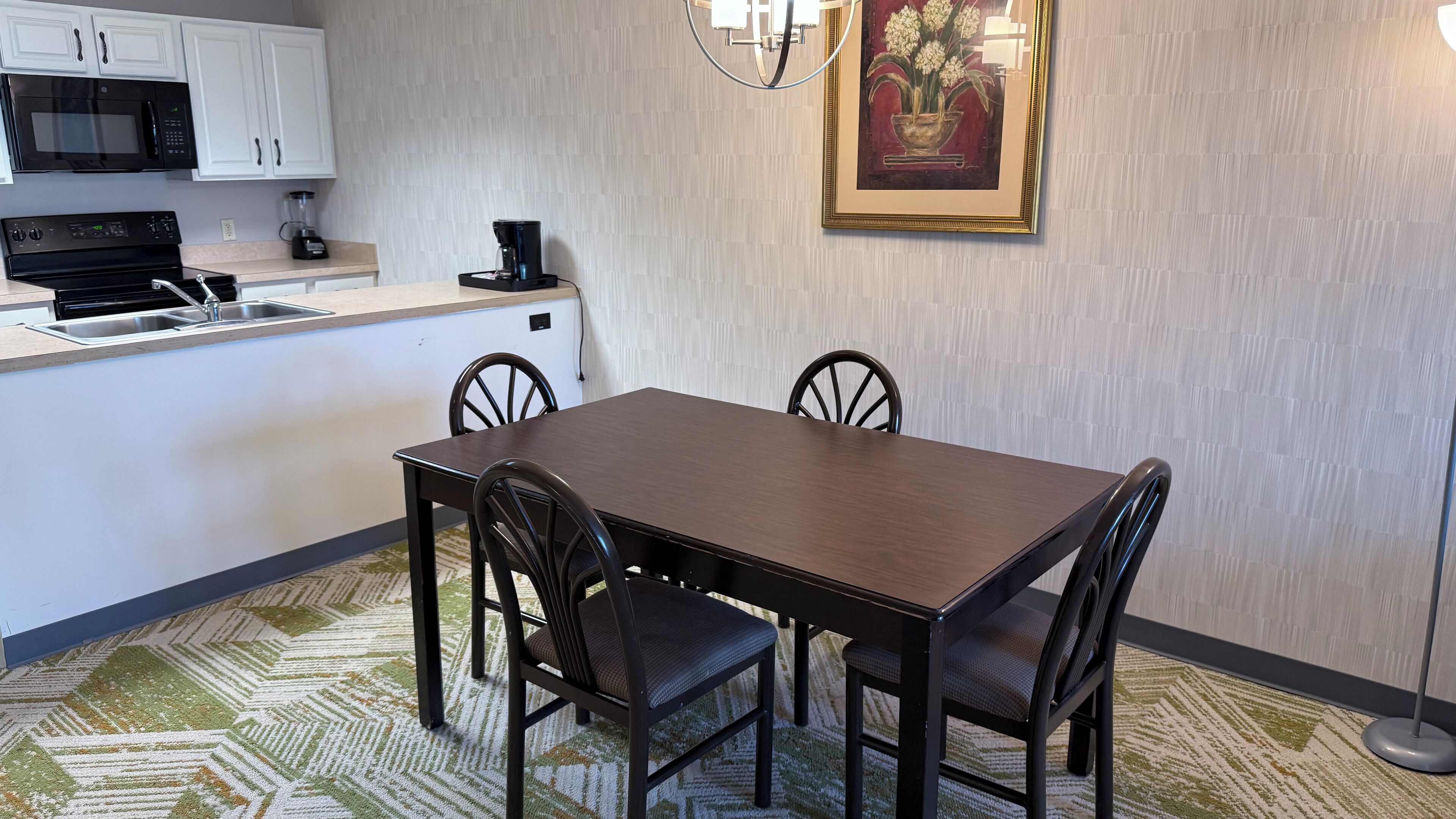 A compact kitchen with a microwave, coffee maker, and a small island. There is a dining table with four chairs and a patterned rug. A framed artwork hangs on the wall.