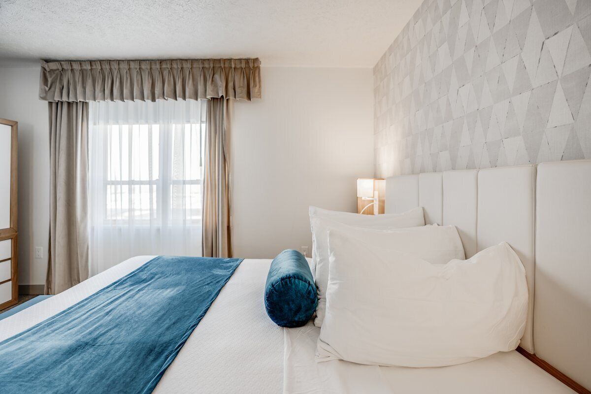 A hotel room with a bed, a blue throw blanket, white pillows, a nightstand with a lamp, and a geometric-patterned wall.