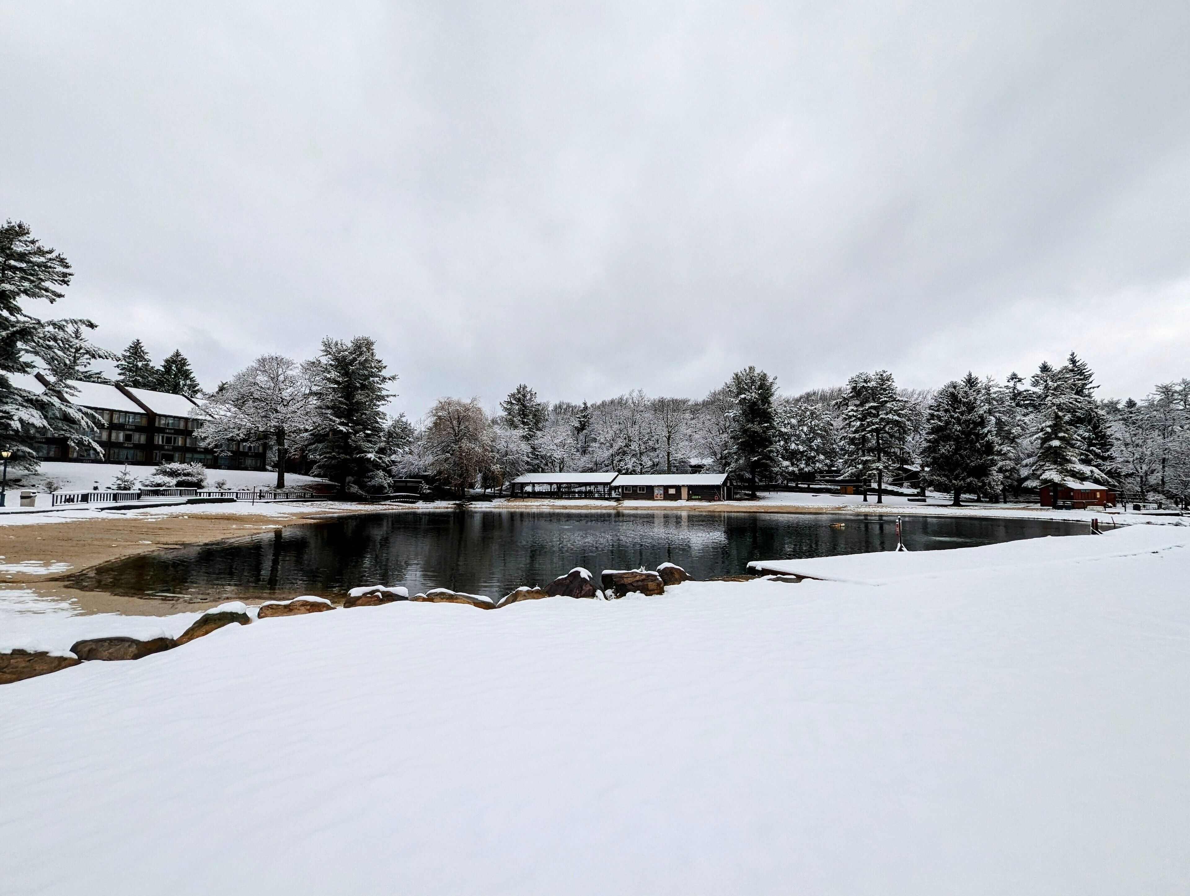 Cena de inverno com paisagem coberta de neve, um lago e edifícios ao fundo.
