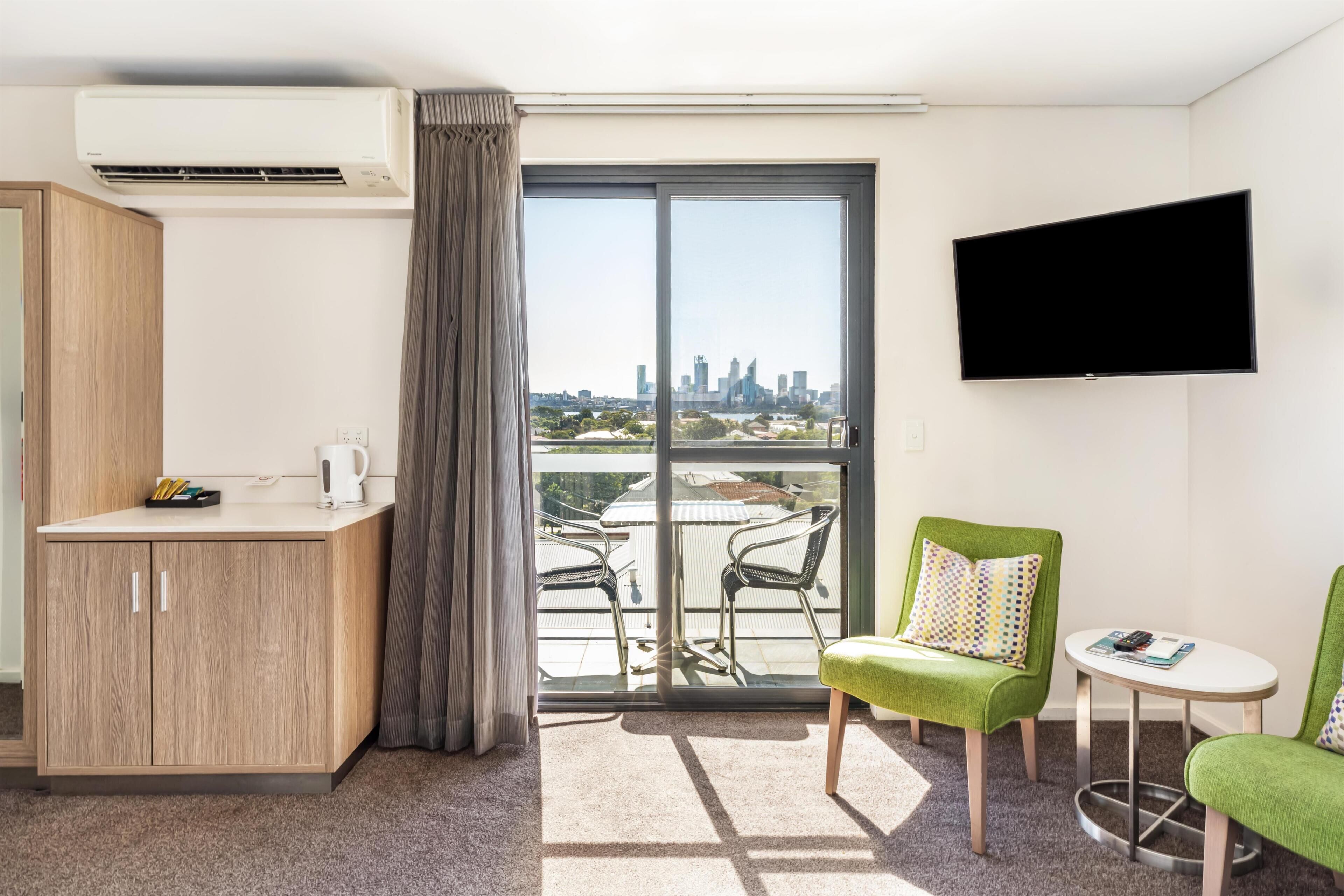 Une chambre d’hôtel avec un balcon, un téléviseur à écran plat, une armoire en bois et deux chaises vertes.
