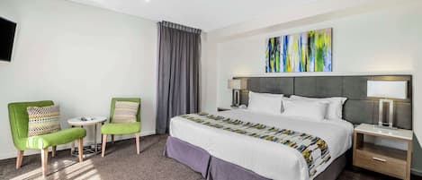 Una habitación de hotel con una cama grande, dos sillas verdes, una mesita y una mesita de noche con lámpara.