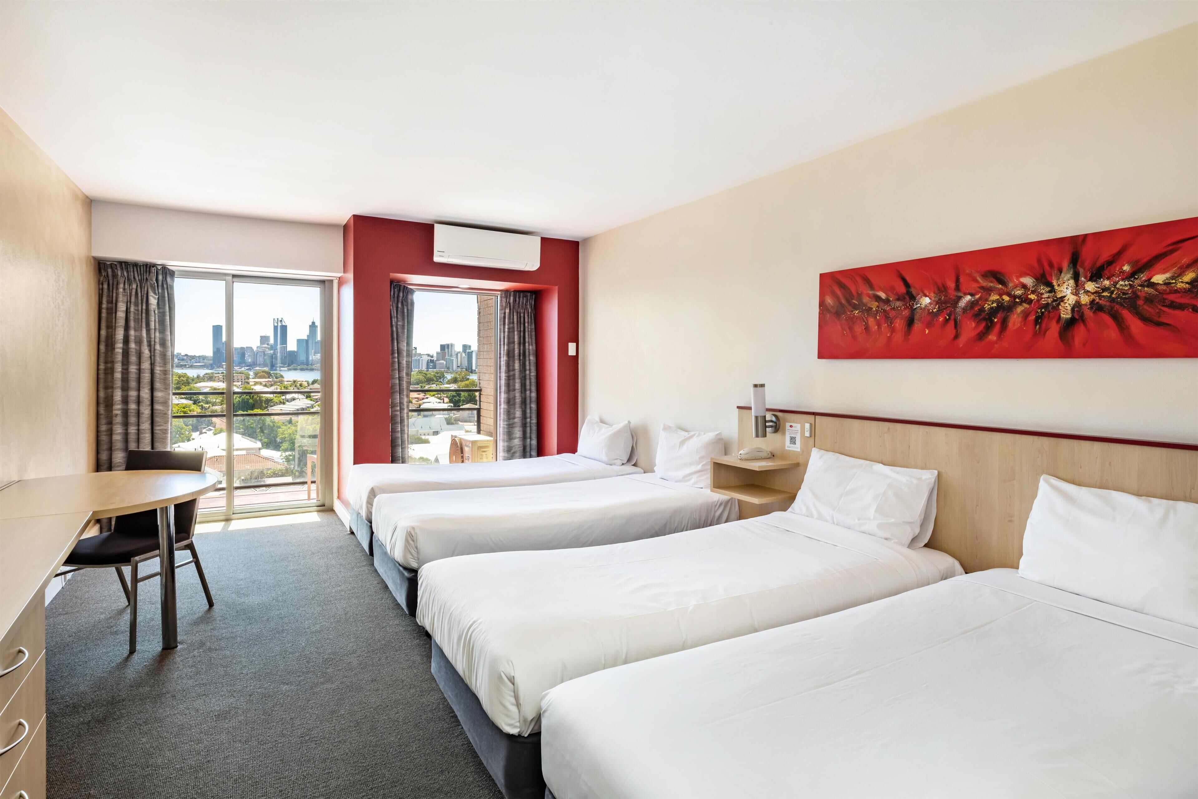 A hotel room with three beds, a desk, a chair, and a large window with a city view.