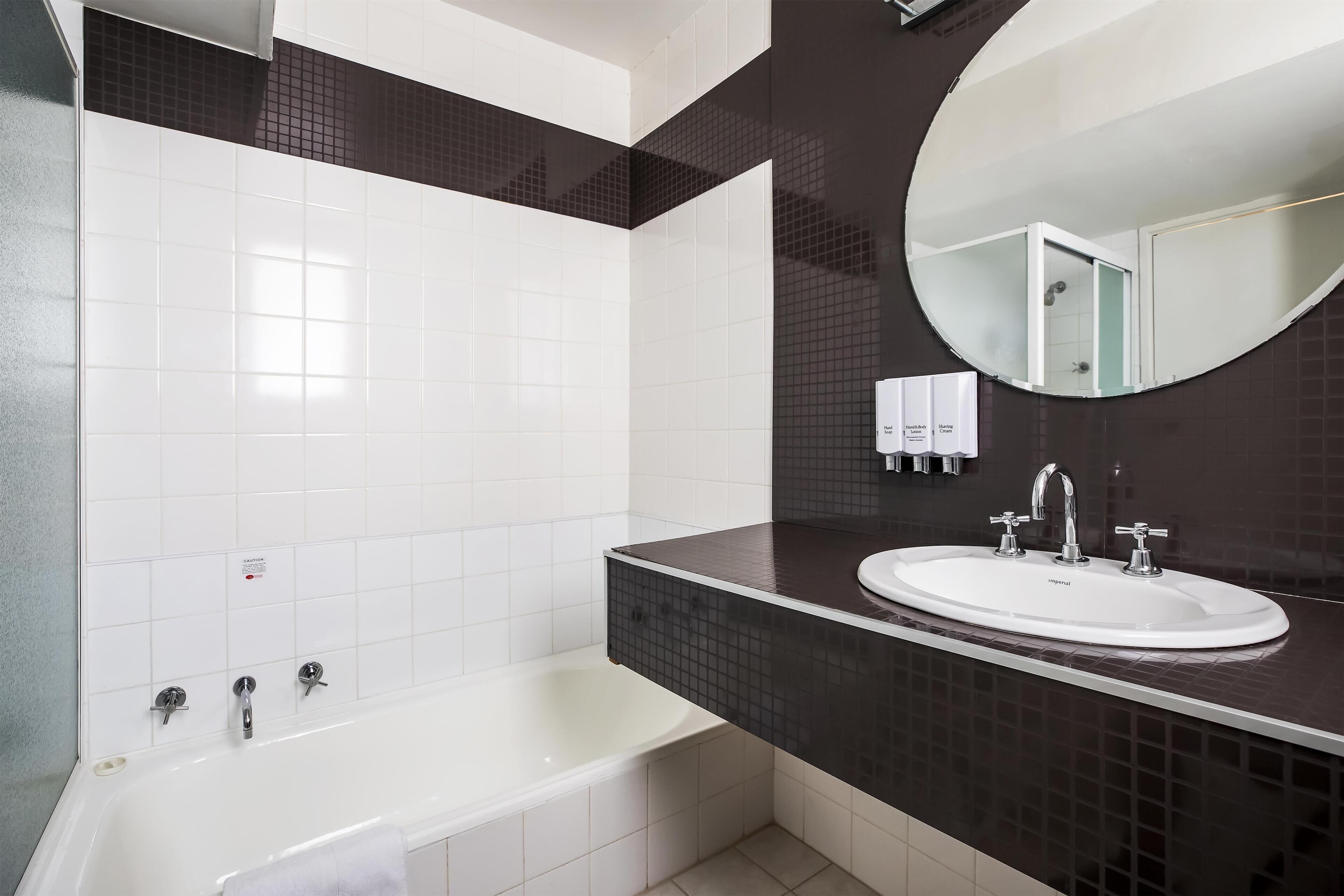 A modern bathroom with a white bathtub, a sink with two faucets, a mirror, and a dark tiled wall.