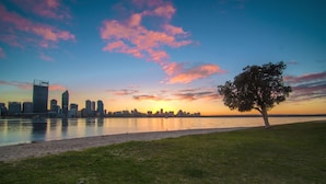 A city skyline at sunset with a river in the foreground and a lone tree on the right. - Metro Hotel Perth (South Perth)