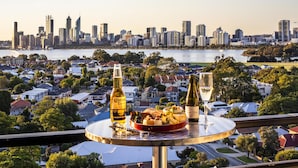 A table with a plate of food, two bottles, and a glass on a balcony overlooking a cityscape and water. - Metro Hotel Perth (South Perth)