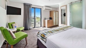 A modern hotel room with a large bed, a green armchair, a small table with a bottle, a TV mounted on the wall, and a sliding door leading to a balcony. - Metro Hotel Perth (South Perth)