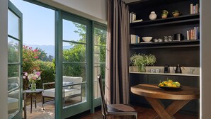 A room with a large sliding glass door leading to a patio, a round wooden table with a bowl of fruit, a bookshelf with books and decorative items, and a chair. - Ojai Valley Inn (Ojai)
