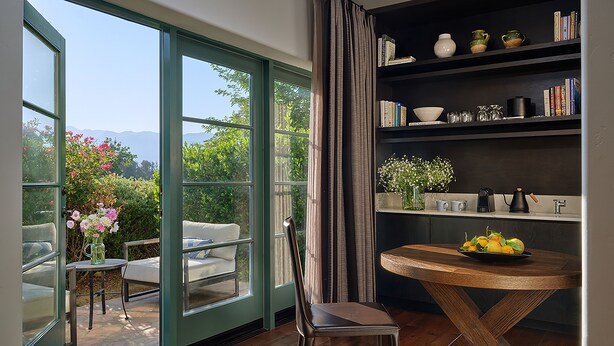 A room with a large sliding glass door leading to a patio, a round wooden table with a bowl of fruit, a bookshelf with books and decorative items, and a chair. - Ojai Valley Inn (Ojai)