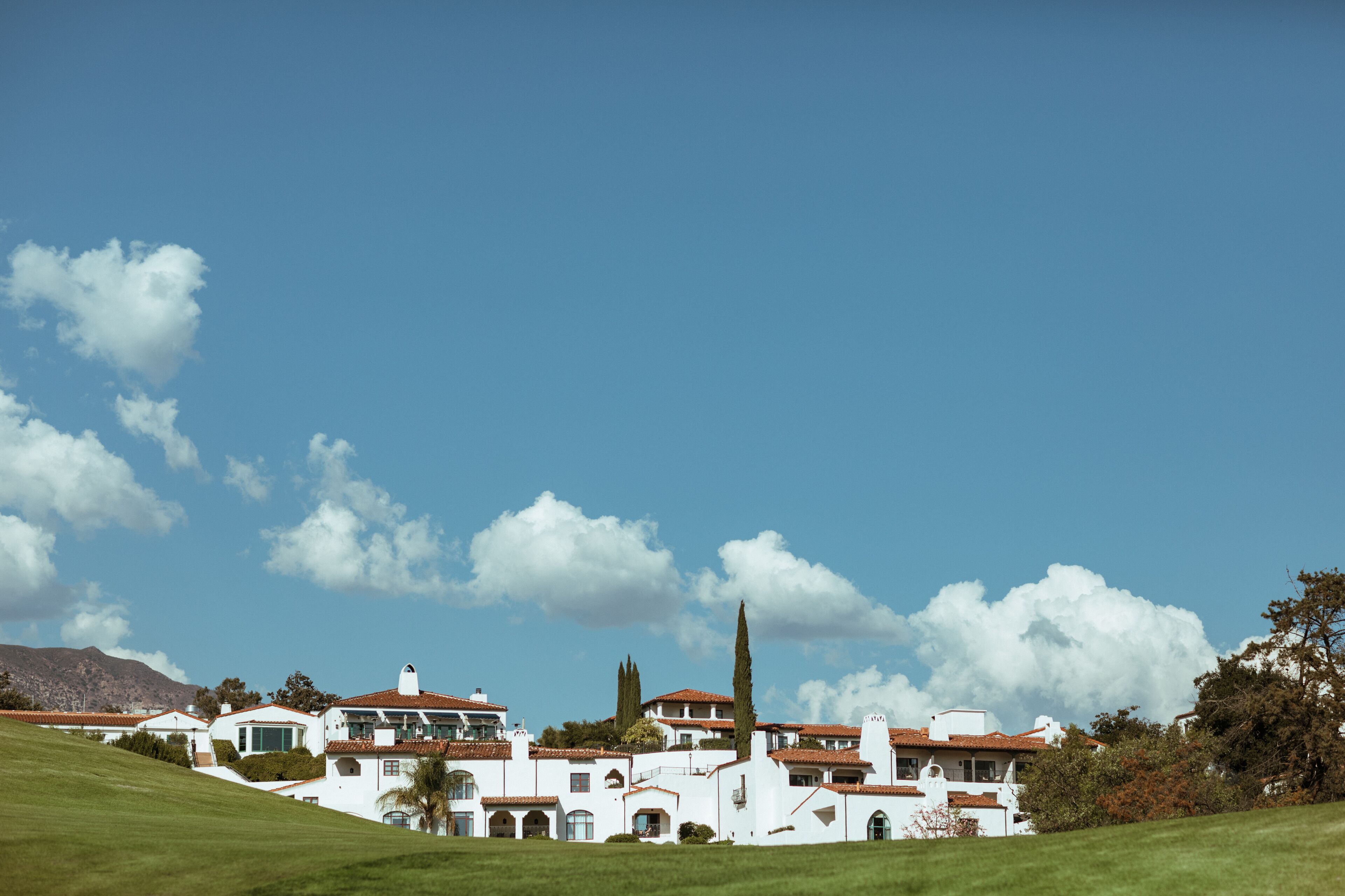 A large white building with multiple levels and a red-tiled roof, surrounded by green grass and trees.