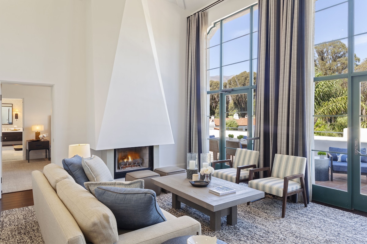 A modern living room with a fireplace, comfortable seating, and large windows offering a view of the outdoors.