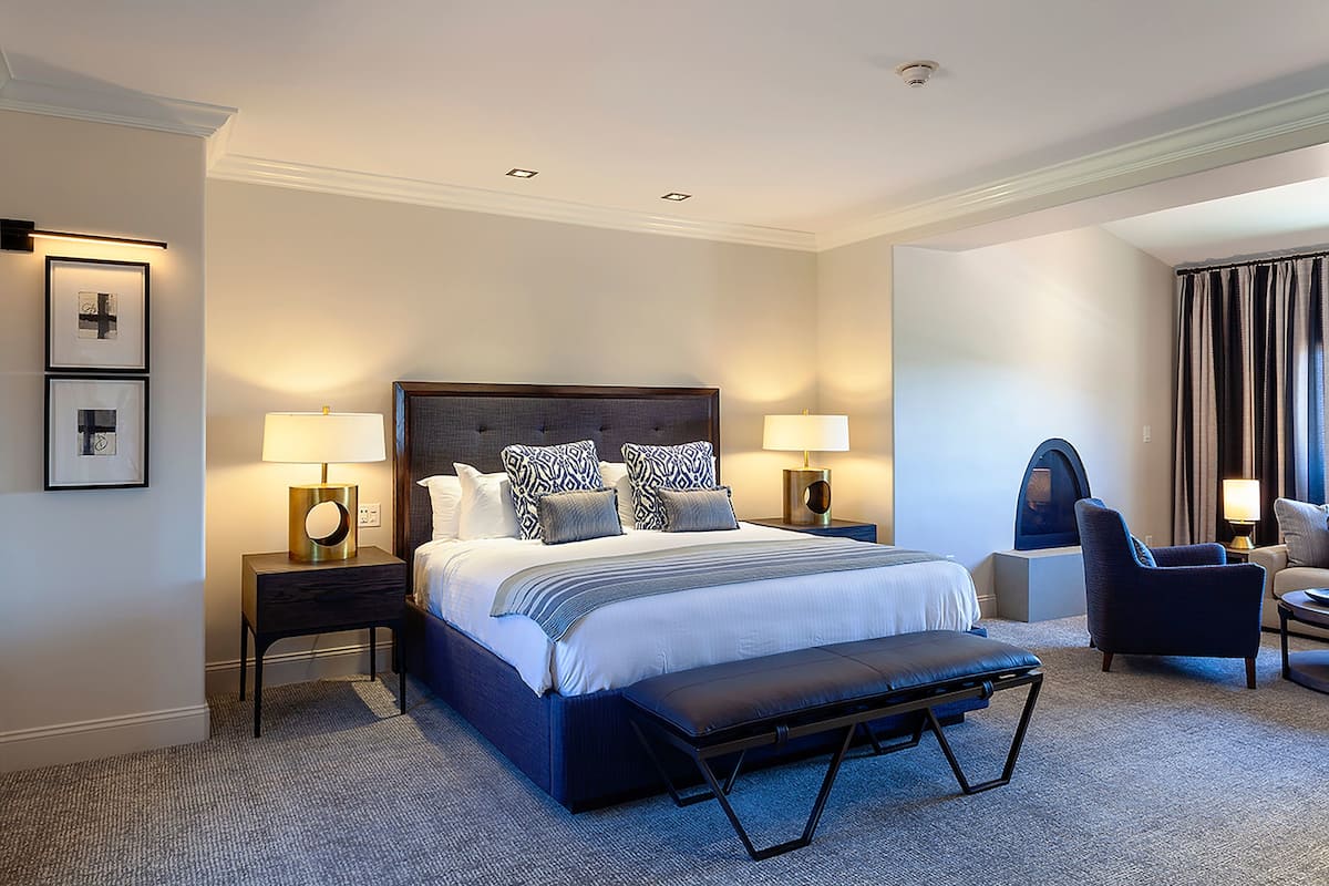 Une chambre d’hôtel avec un grand lit, deux tables de chevet avec des lampes, un banc, une chaise et un canapé.
