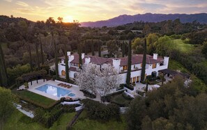 A large, white, two-story house with a swimming pool, surrounded by trees and mountains.