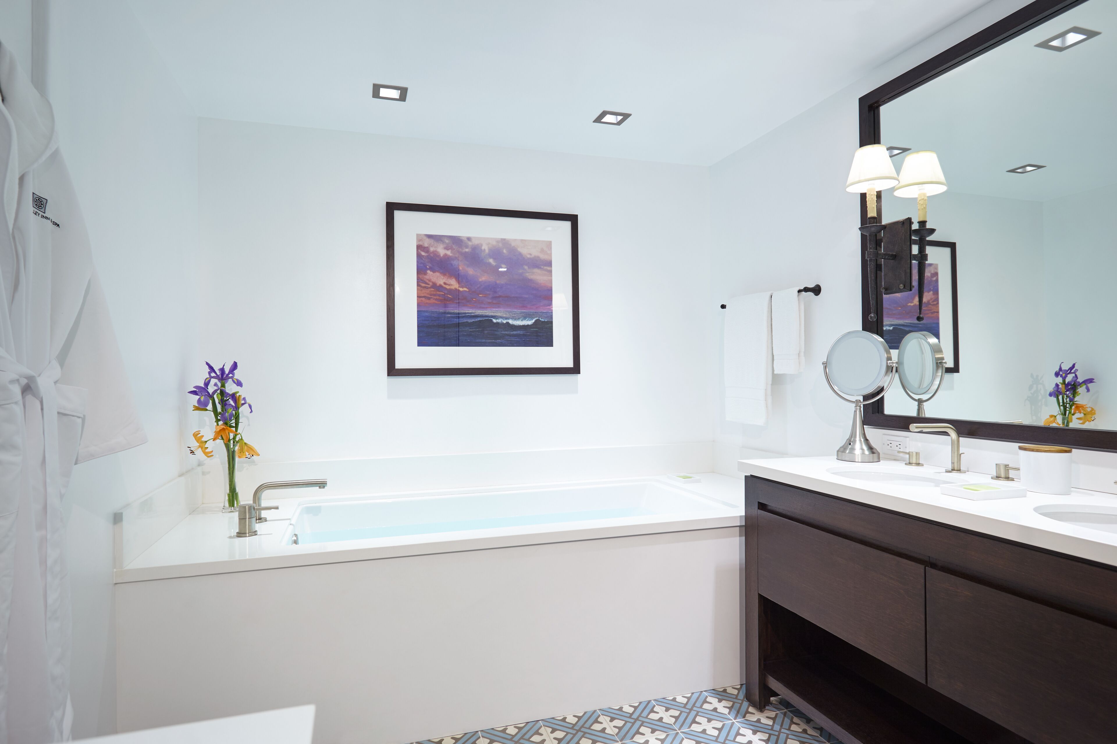 Une salle de bain moderne avec une baignoire, deux lavabos, un grand miroir et un tableau représentant un coucher de soleil sur l’océan.