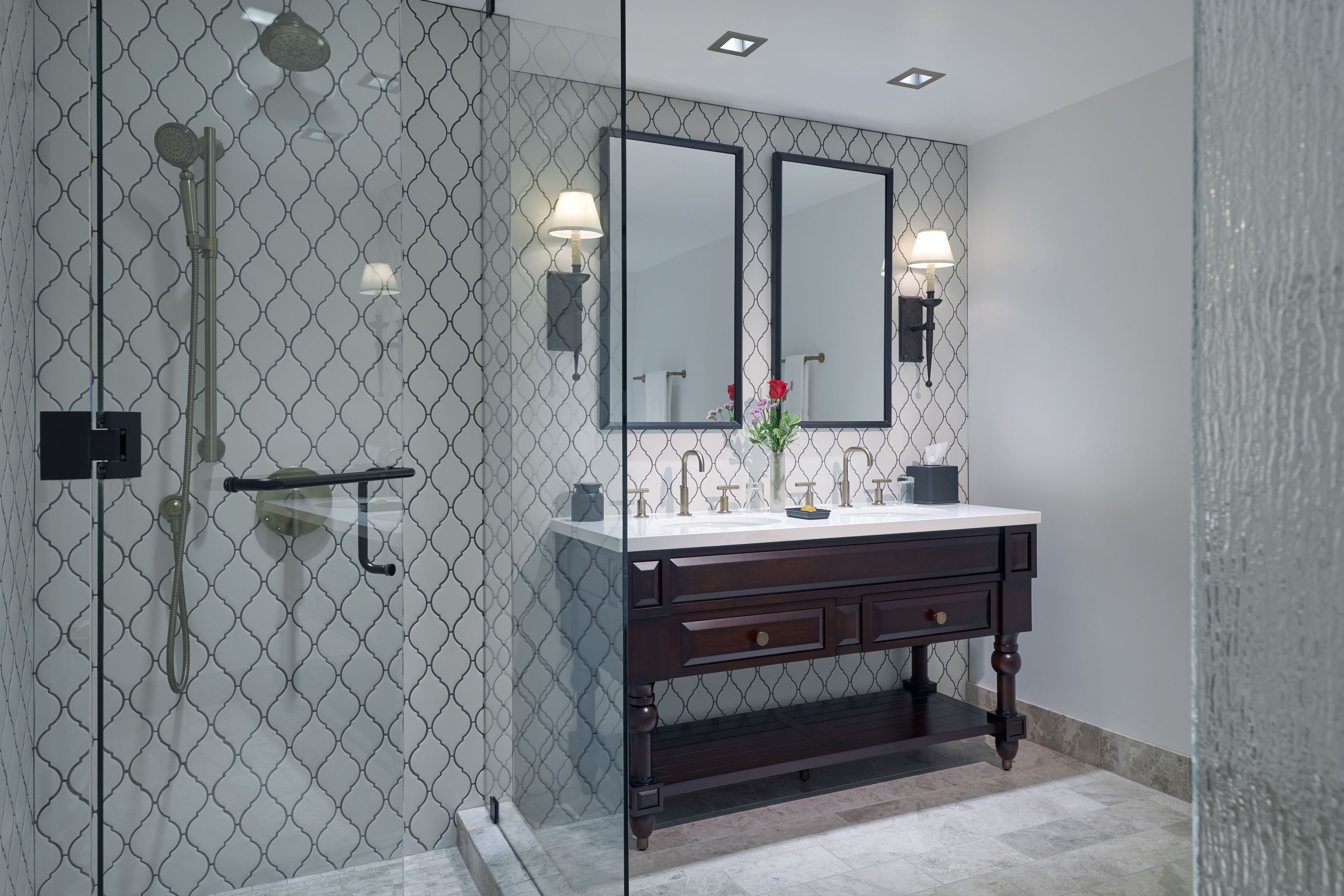 A modern bathroom with a glass-enclosed shower, a double vanity with two mirrors, and a small vase with flowers on the vanity.