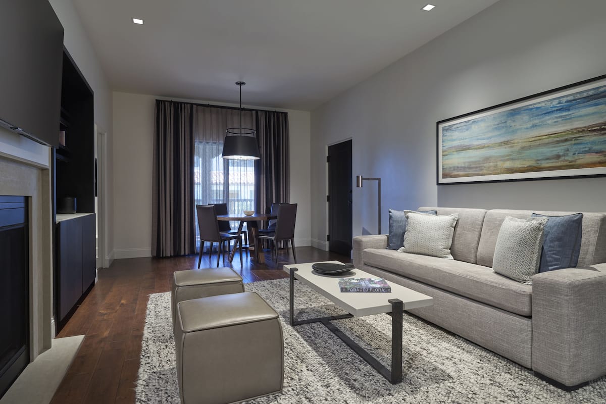 Un salon moderne comprenant un canapé, une table basse et un pouf. On y trouve également un coin repas avec une table et des chaises, ainsi qu’une cuisine équipée de placards et d’un réfrigérateur.