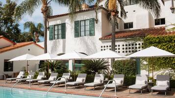 A poolside area with lounge chairs and umbrellas, palm trees, and a building with green shutters.