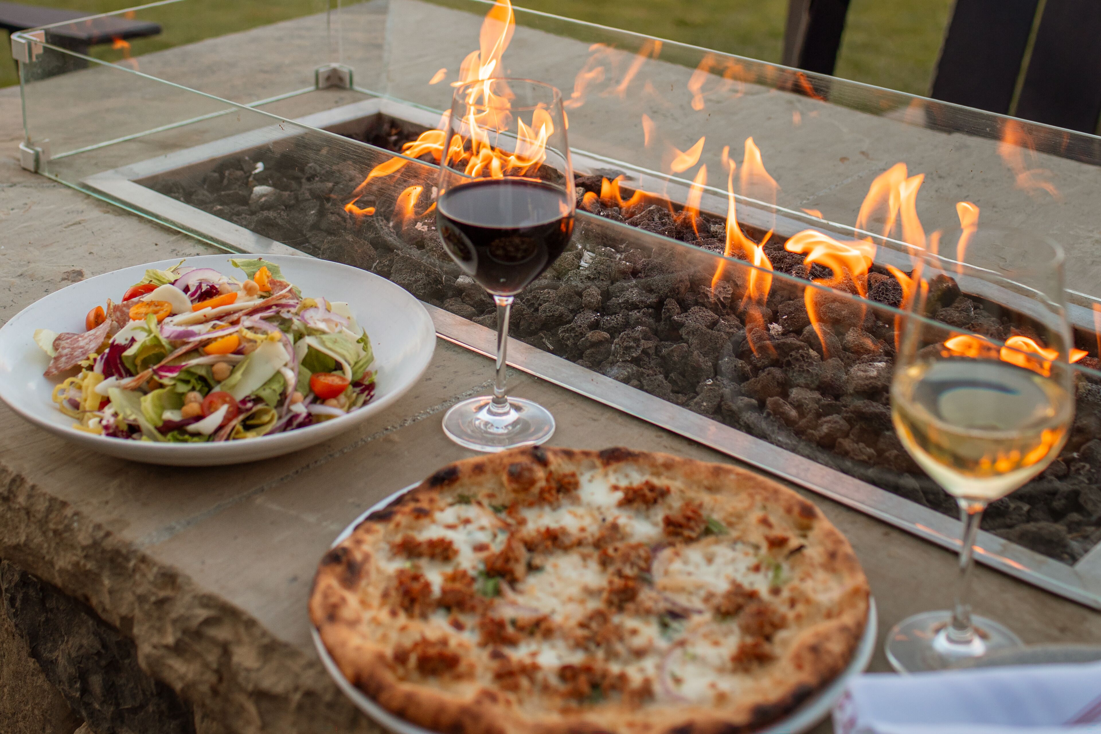 A glass-top table with a fire feature, two glasses of wine, a plate of salad, and a pizza.