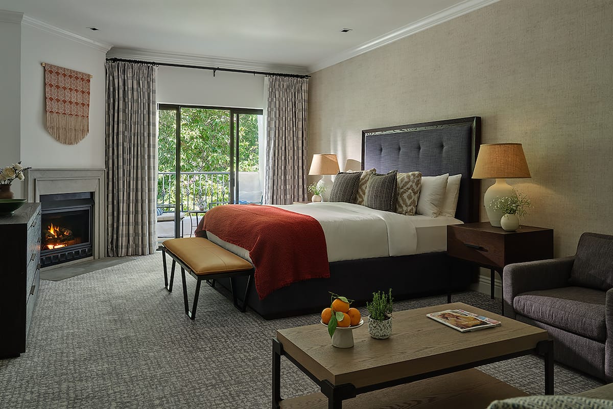 Une chambre d’hôtel avec un lit, des lampes de chevet, une table basse avec des fruits, un banc, une cheminée et une vue sur la verdure à l’extérieur.