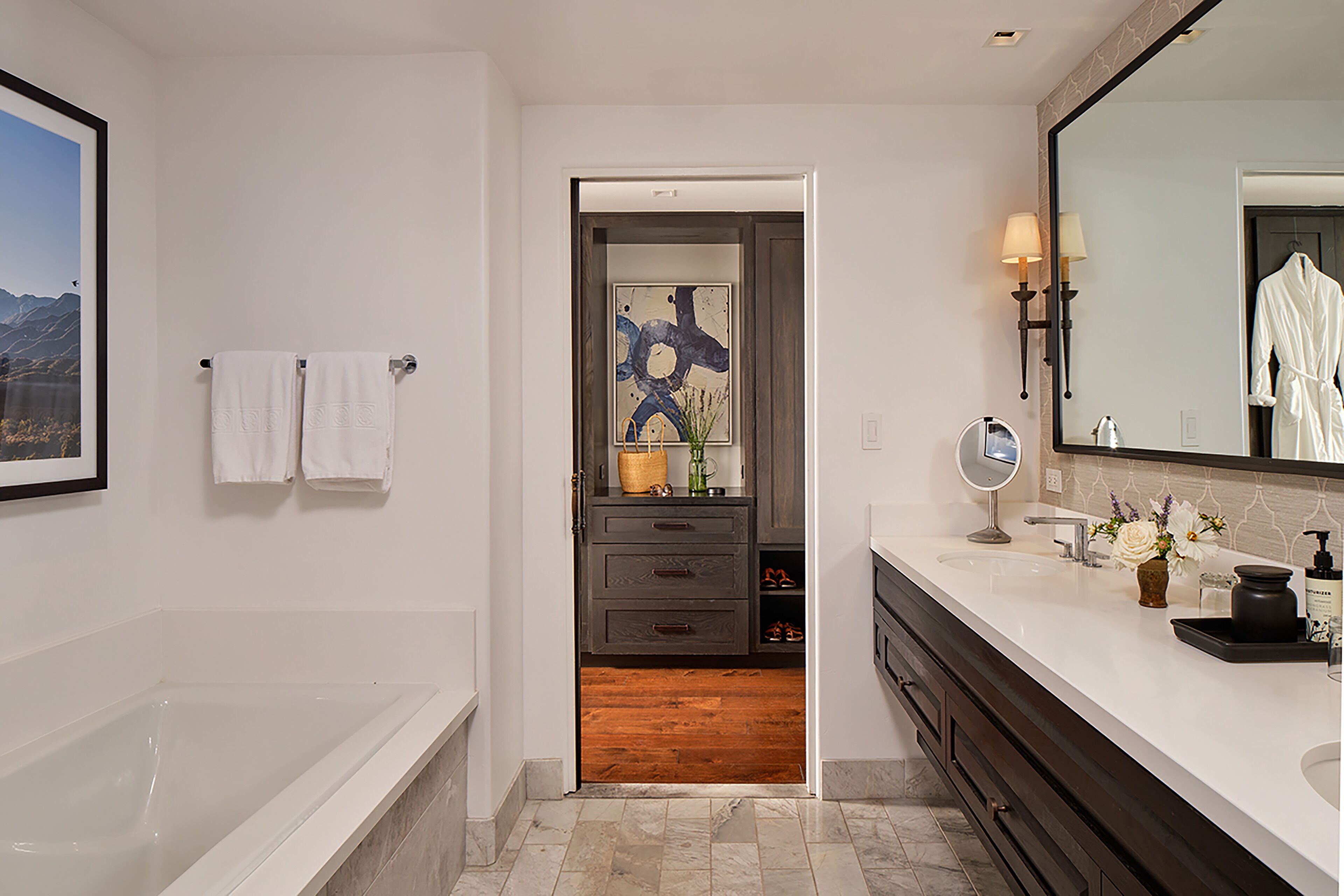 Un baño moderno con bañera, tocador con espejo y suelo de madera que conduce a un dormitorio.