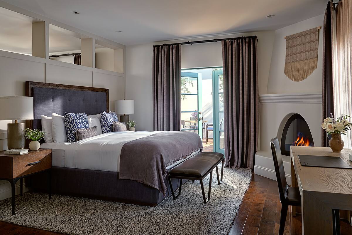 Une chambre moderne avec un grand lit, des tables de chevet, un banc, une cheminée et une vue sur l’extérieur.