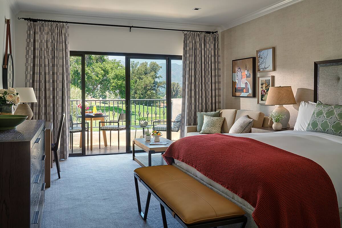 Une chambre d’hôtel avec un grand lit, un banc, une table ornée d’un vase de fleurs et un balcon offrant une vue sur la verdure et les montagnes.