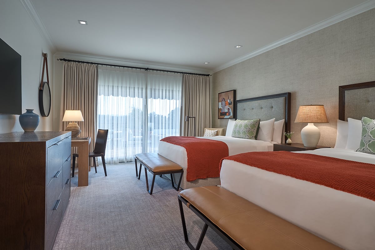 Une chambre d’hôtel avec un lit, un bureau, une chaise, un banc, une télévision et une fenêtre avec des rideaux.