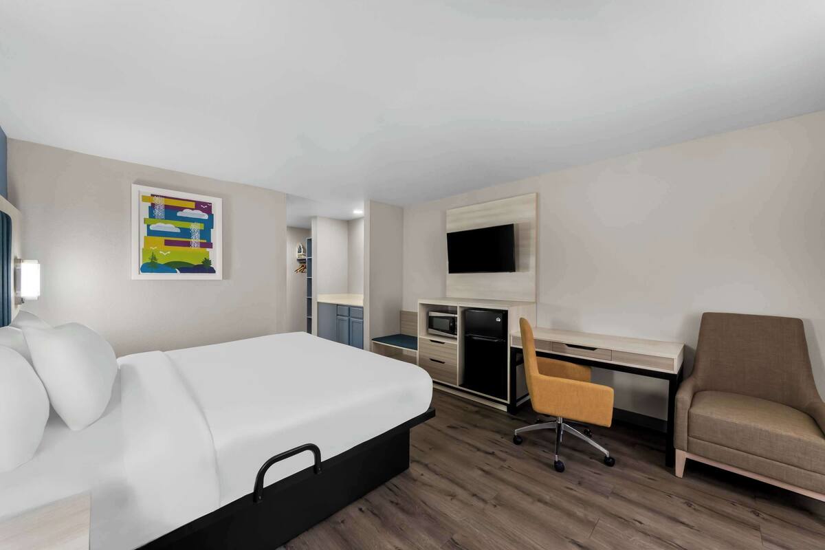 A hotel room with a bed, desk, chair, TV, and kitchenette.