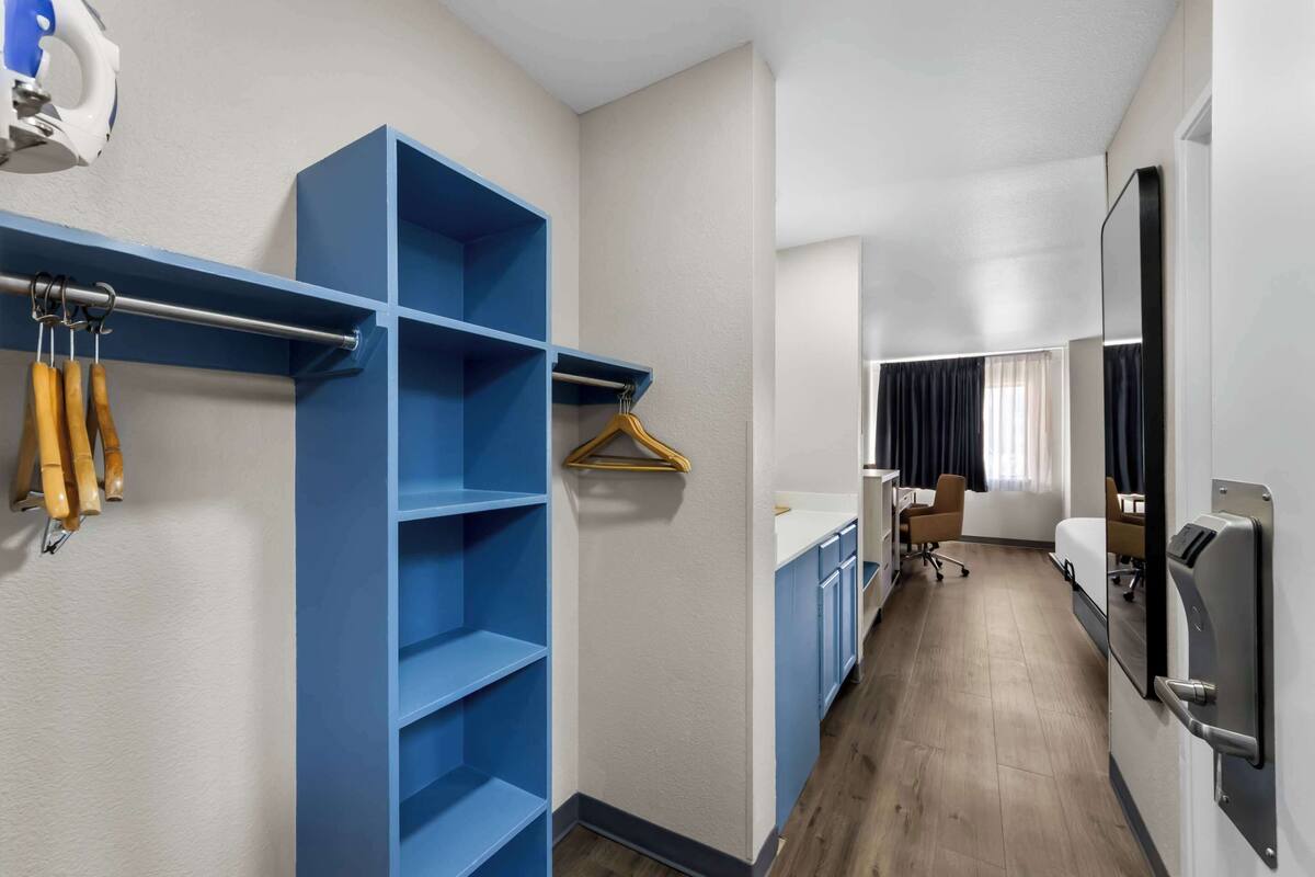A hotel room with a blue shelving unit, a wooden coat rack, a desk with a chair, and a large mirror.