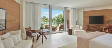 A modern hotel room with a large bed, a sofa, a dining table with chairs, a TV, and a balcony with a view of the ocean.