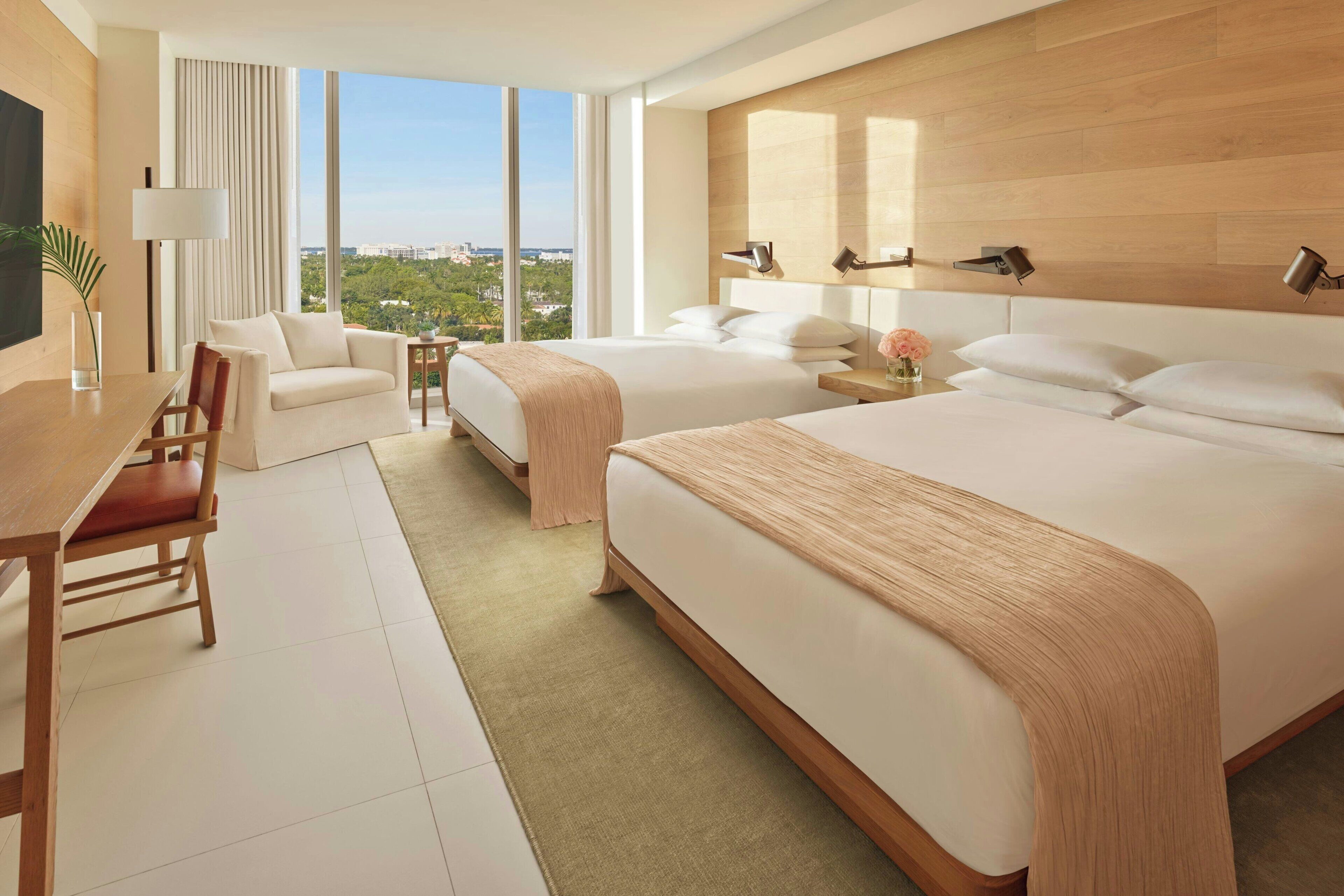 A modern hotel room with two beds, a desk, and a view of the city.