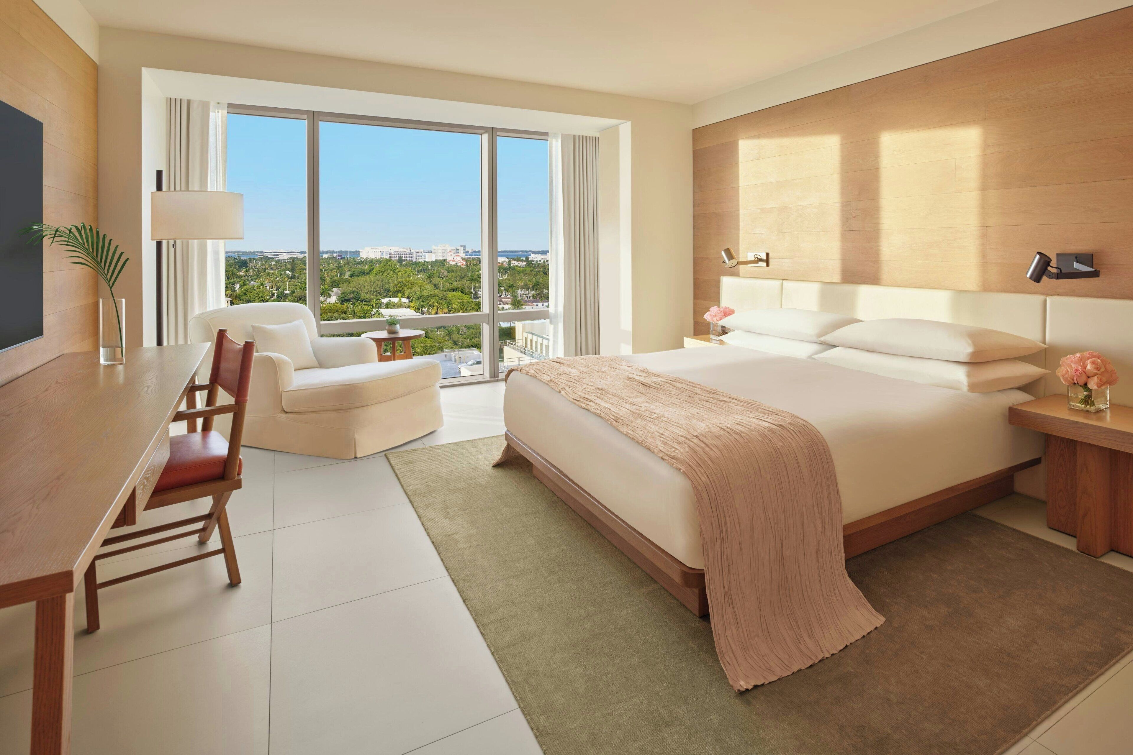 A modern hotel room with a large bed, a desk, a chair, and a view of the city through large windows.